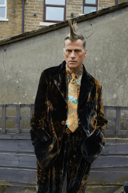 A man with a mohawk stands outdoors wearing a velvet suit and patterned tie, posing against a weathered wall and a glimpse of a building.