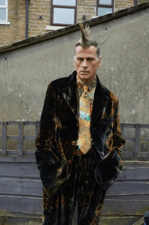 A man with a mohawk stands outdoors wearing a velvet suit and patterned tie, posing against a weathered wall and a glimpse of a building.