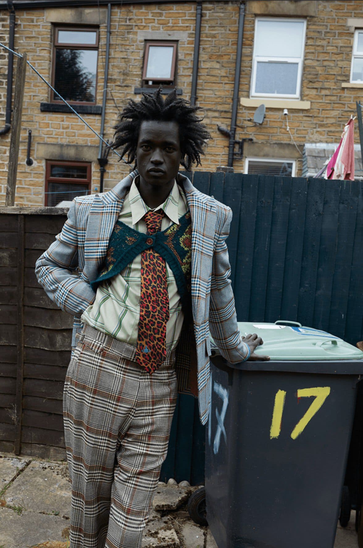 Portrait of a Black male in a plaid suit leaning against a trash bin in an urban backyard, with a brick building in the background.