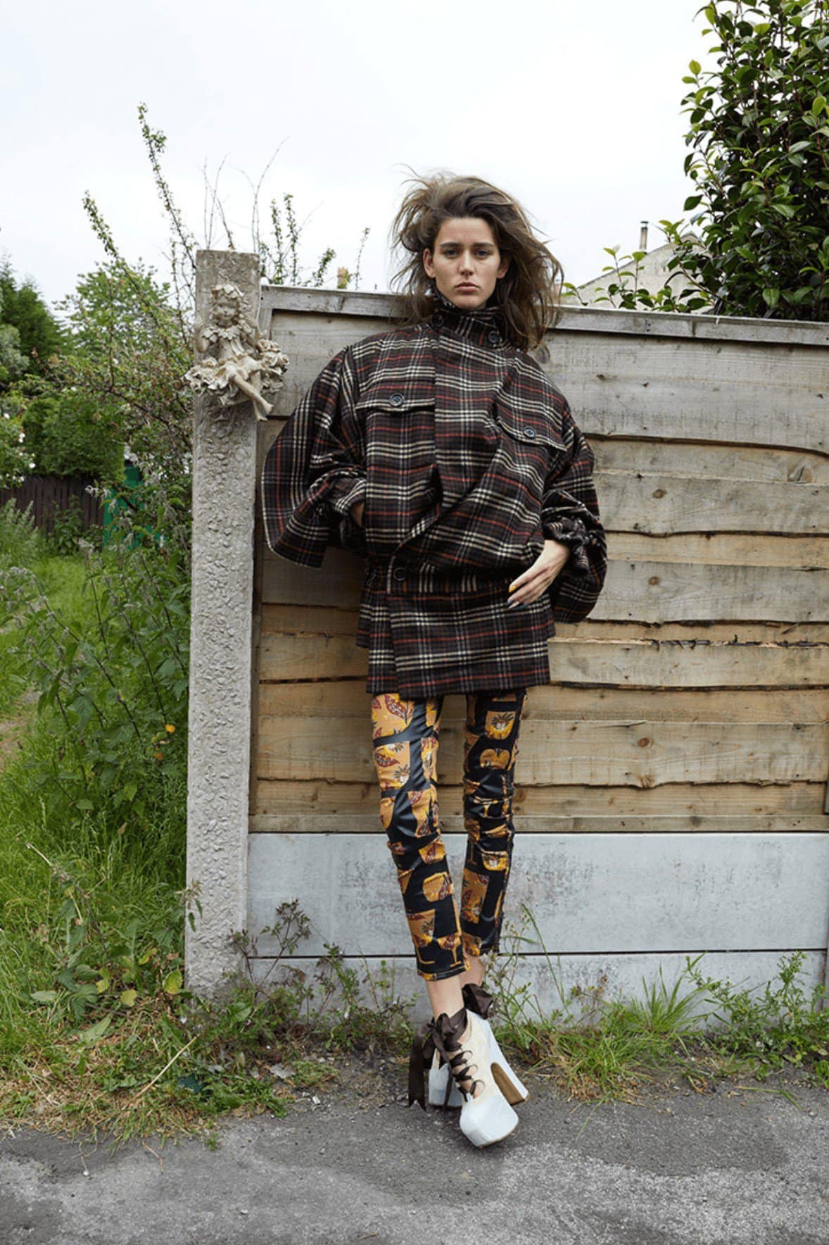 A woman stands in an outdoor setting, wearing a plaid jacket, patterned leggings, and white platform boots against a wooden fence.