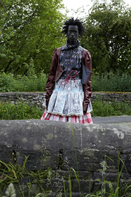 Adult fashion model in outdoor setting. Dark-skinned individual wears a mixed-fabric outfit with a brown leather jacket and frilly skirt.