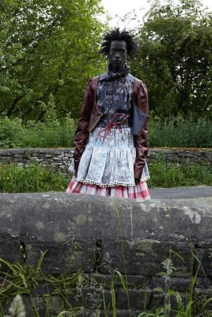 Adult fashion model in outdoor setting. Dark-skinned individual wears a mixed-fabric outfit with a brown leather jacket and frilly skirt.