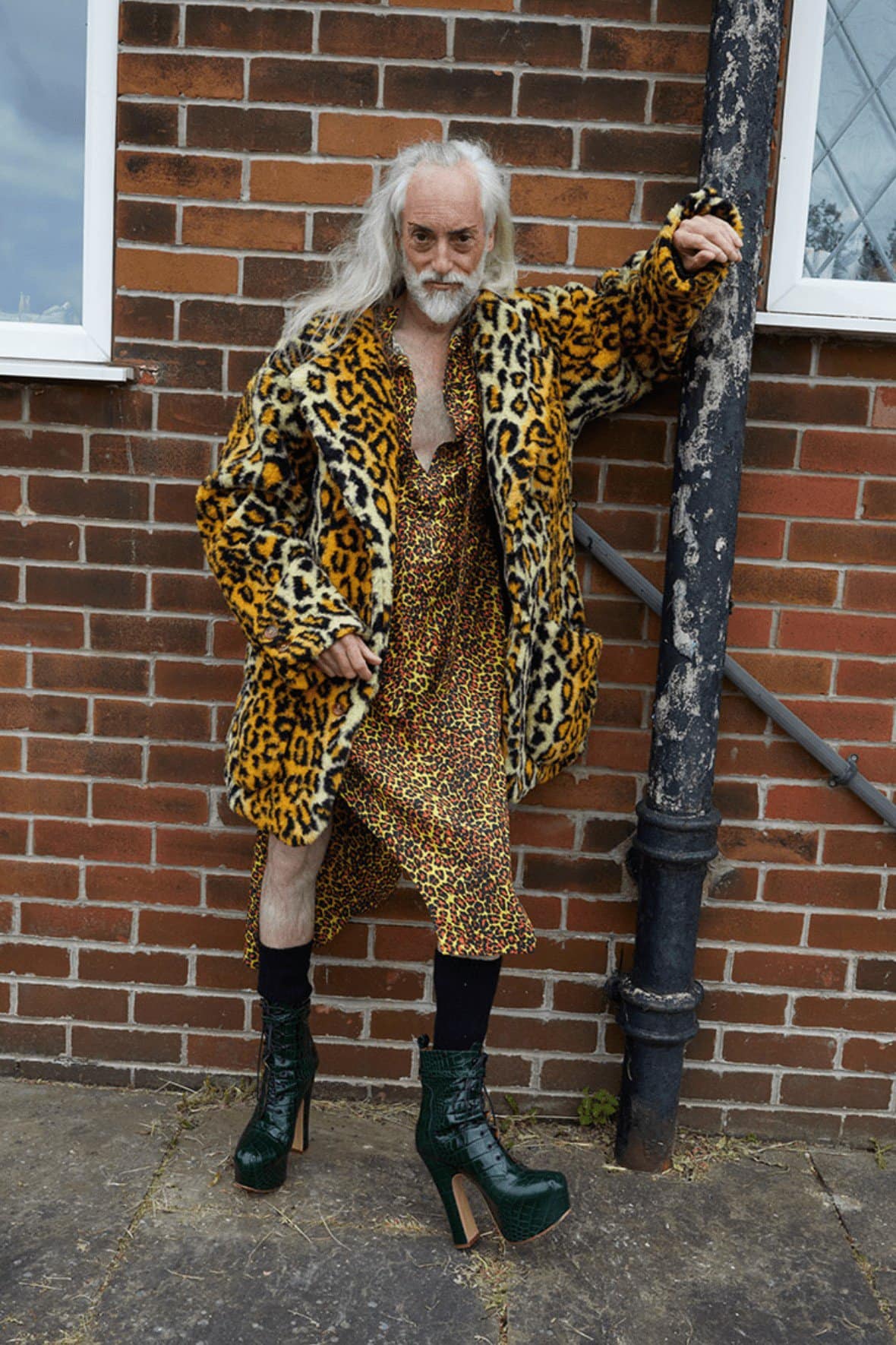 A person with long hair and a beard poses in a leopard-print coat and dress outside against a brick wall, accessorized with dark socks and green platform boots.