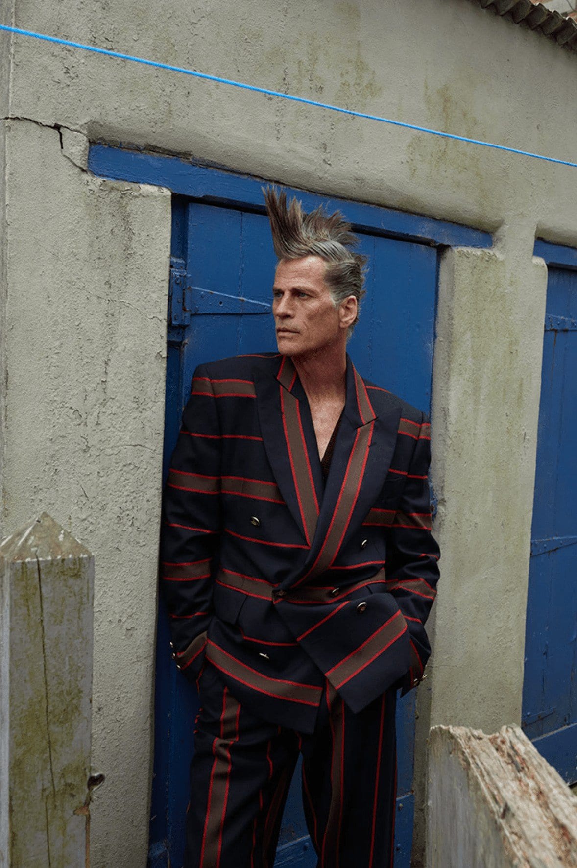 Stylish older man with a mohawk, wearing a striped suit, poses against a blue-doored building in an outdoor setting. Fashion editorial.