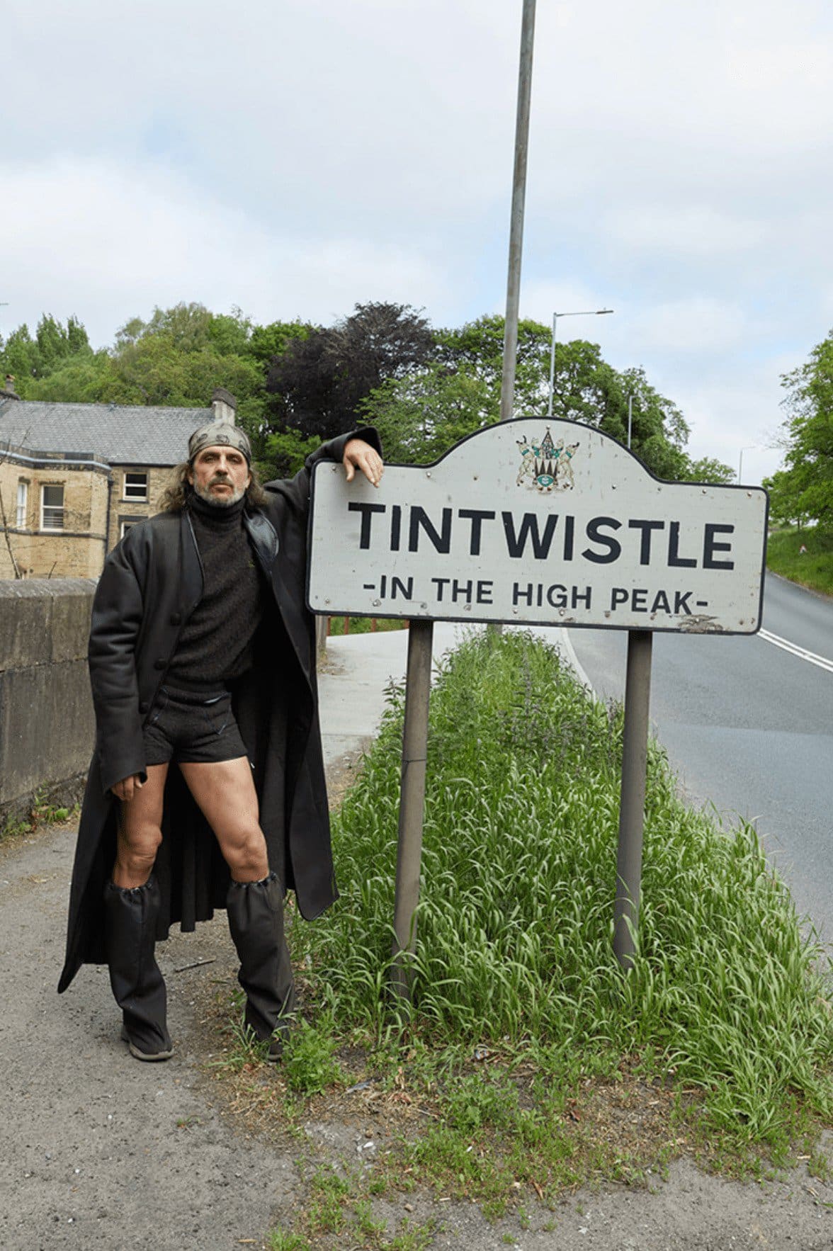 Man in medieval-style costume poses by Tintwistle village sign on a rural road with green grass and foliage in the background on an overcast day.