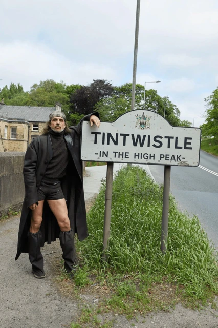 Man in medieval-style costume poses by Tintwistle village sign on a rural road with green grass and foliage in the background on an overcast day.