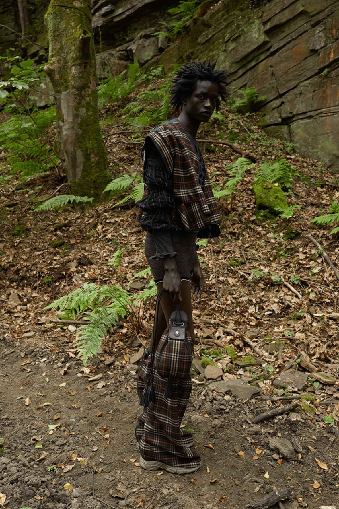Adult male fashion model in plaid clothing poses on dirt path in a forest setting with trees, plants, and rock formations.
