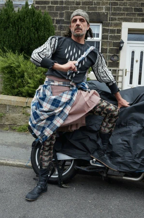 Man wearing a patterned shirt, plaid skirt, and headscarf standing beside a covered motorcycle outdoors.