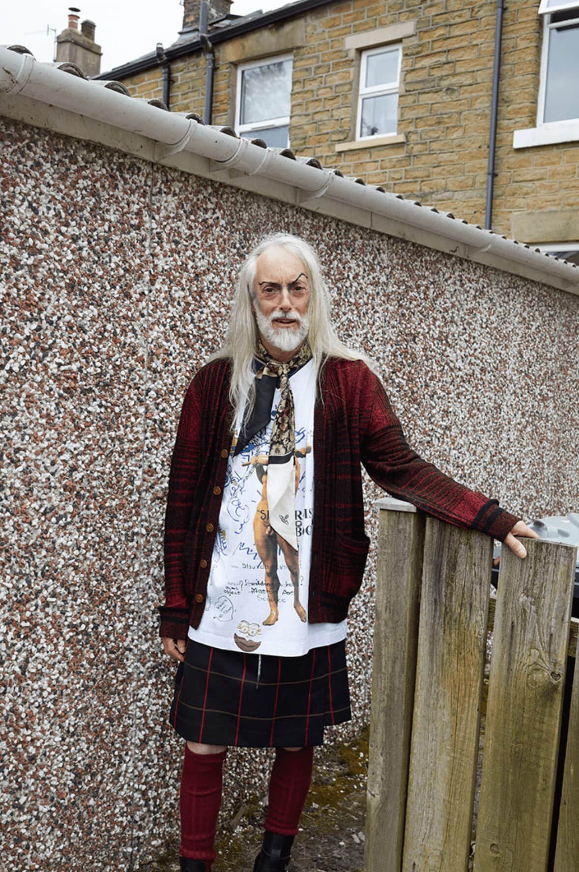 Mature adult wearing kilt, patterned cardigan, t-shirt with print, and scarf posing outdoors in front of wall near fence and building.