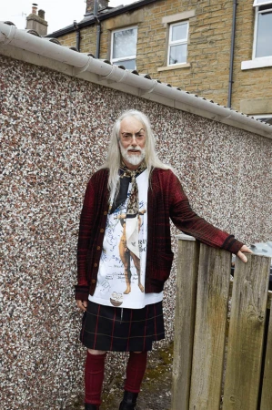 Mature adult wearing kilt, patterned cardigan, t-shirt with print, and scarf posing outdoors in front of wall near fence and building.