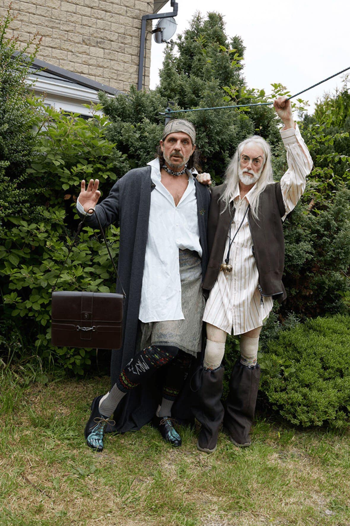 Two adults dressed in theatrical costumes pose outdoors; one carries a briefcase, and both sport unique footwear amidst green foliage.