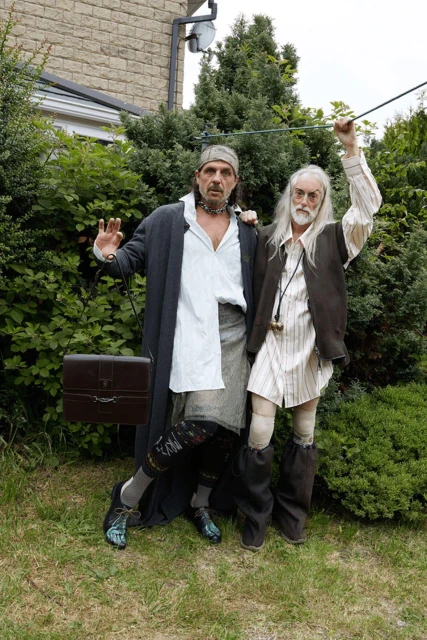 Two adults dressed in theatrical costumes pose outdoors; one carries a briefcase, and both sport unique footwear amidst green foliage.