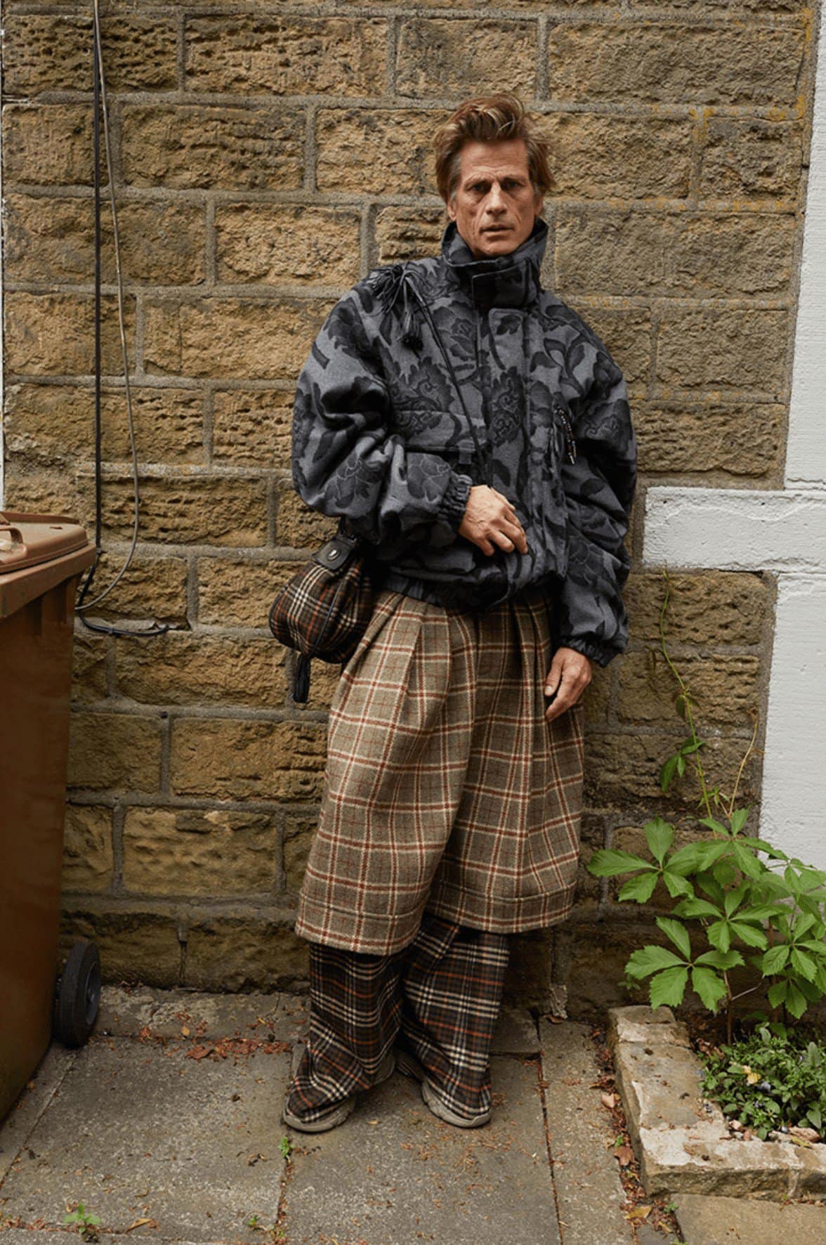 Adult male poses in grey floral print puffer jacket, plaid skirt-pants, and tan shoulder bag against a brick wall in an outdoor setting.