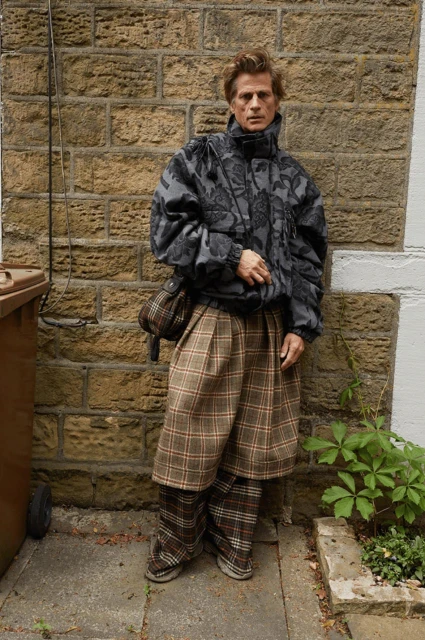 Adult male poses in grey floral print puffer jacket, plaid skirt-pants, and tan shoulder bag against a brick wall in an outdoor setting.