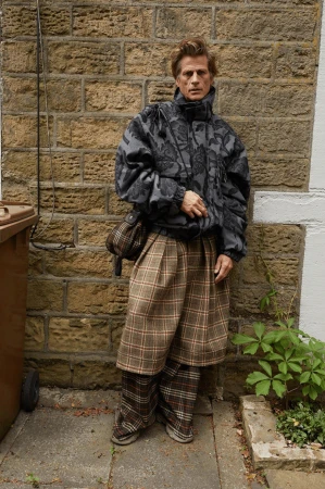 Adult male poses in grey floral print puffer jacket, plaid skirt-pants, and tan shoulder bag against a brick wall in an outdoor setting.