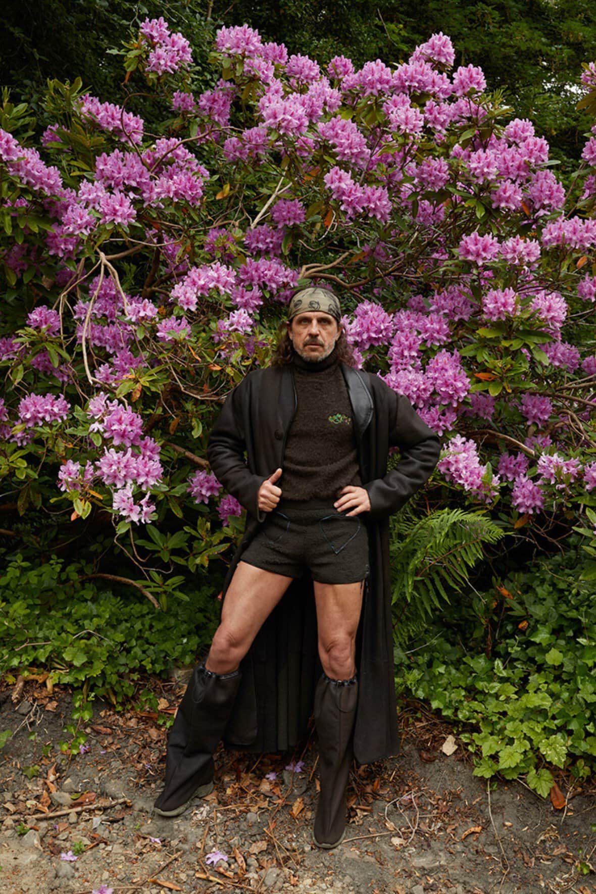 Man in shorts and boots poses in front of blooming rhododendron bush with pink flowers in an outdoor garden setting.