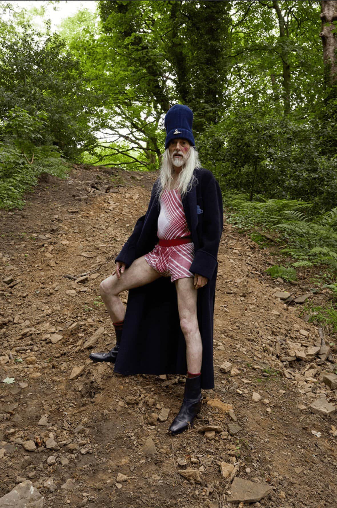 Elderly man in a swimsuit and coat poses on a dirt path outdoors surrounded by trees and greenery in a natural, wooded environment.