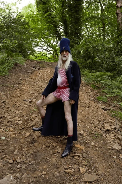 Elderly man in a swimsuit and coat poses on a dirt path outdoors surrounded by trees and greenery in a natural, wooded environment.