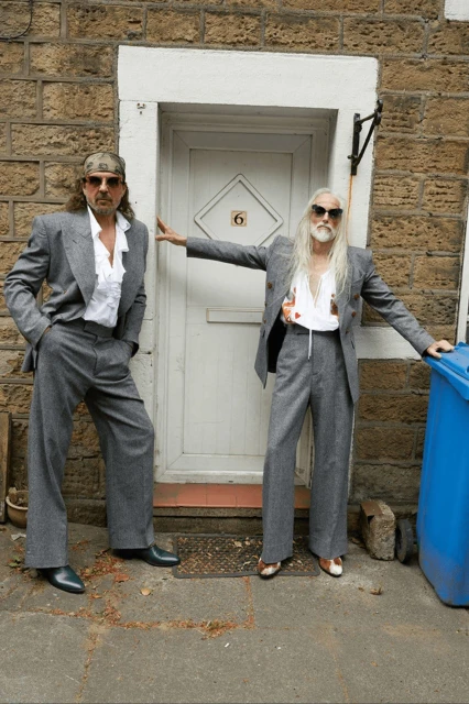 Two adult males pose in grey suits and sunglasses outside a brick building with a white door and blue bin. Fashion editorial shot.