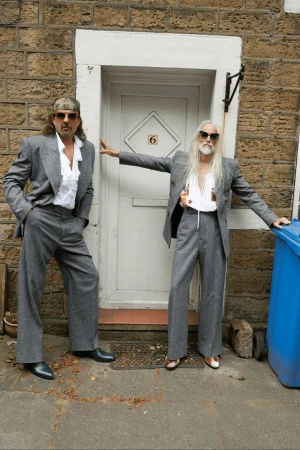 Two adult males pose in grey suits and sunglasses outside a brick building with a white door and blue bin. Fashion editorial shot.