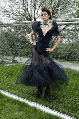 Woman in a black tulle gown stands in front of a soccer net on a grassy field wearing jewelry in a fashion editorial shot.