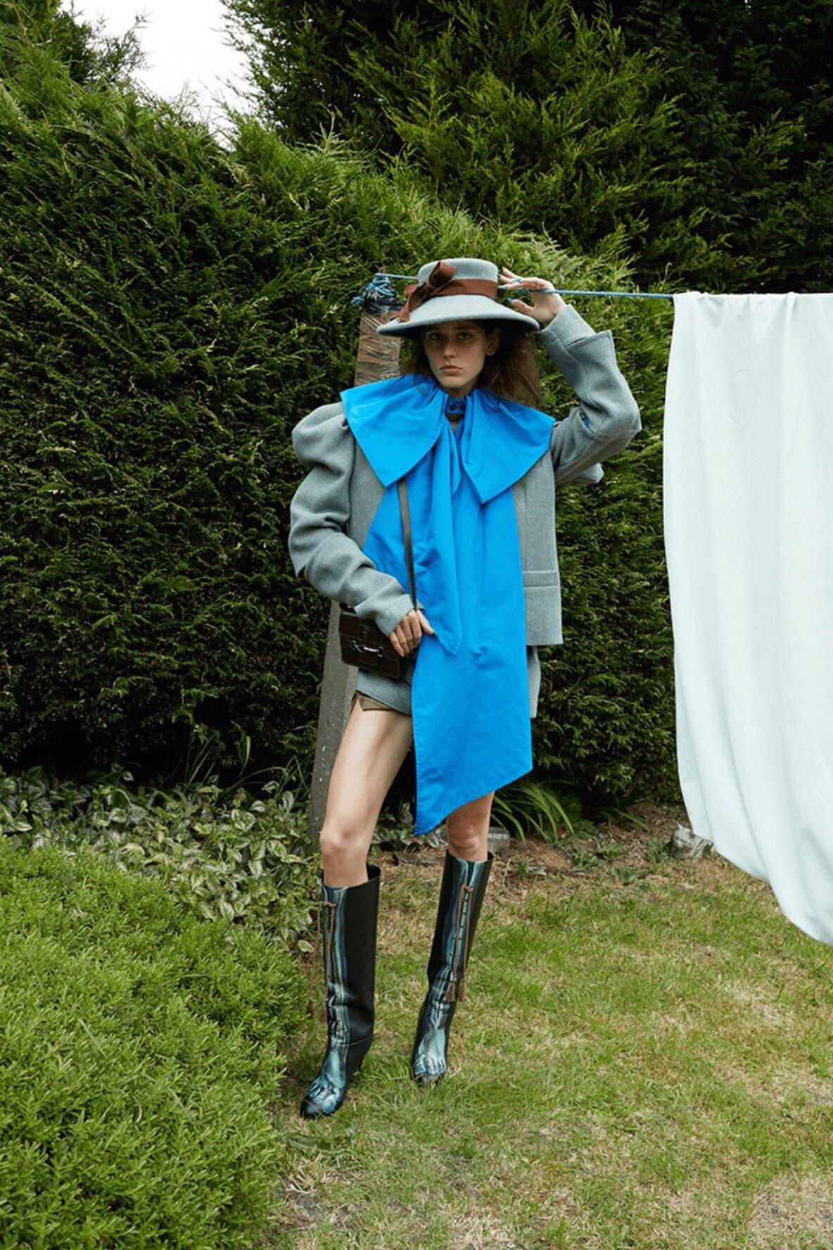 Fashionable woman poses in garden wearing hat, blue scarf, jacket, shorts, shiny boots, and holding purse in outdoor fashion shot.