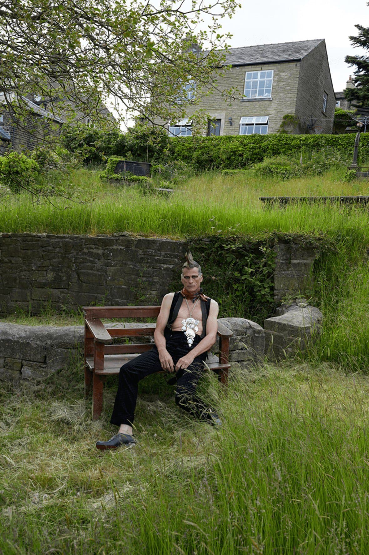 Man with a mohawk sits on a bench in a grassy yard wearing a vest and dark pants, house and greenery in the background.