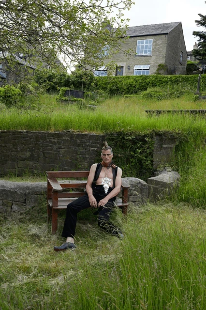 Man with a mohawk sits on a bench in a grassy yard wearing a vest and dark pants, house and greenery in the background.