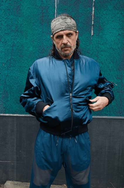 Full-length shot of an adult male with a grey patterned headscarf wearing a shiny dark teal tracksuit against a blue wall.