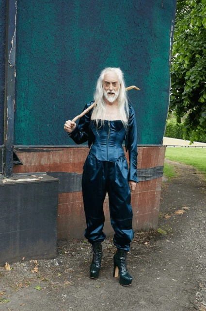 Person in blue satin outfit and platform boots holds stick in outdoor setting with green background and dark structure to the left.