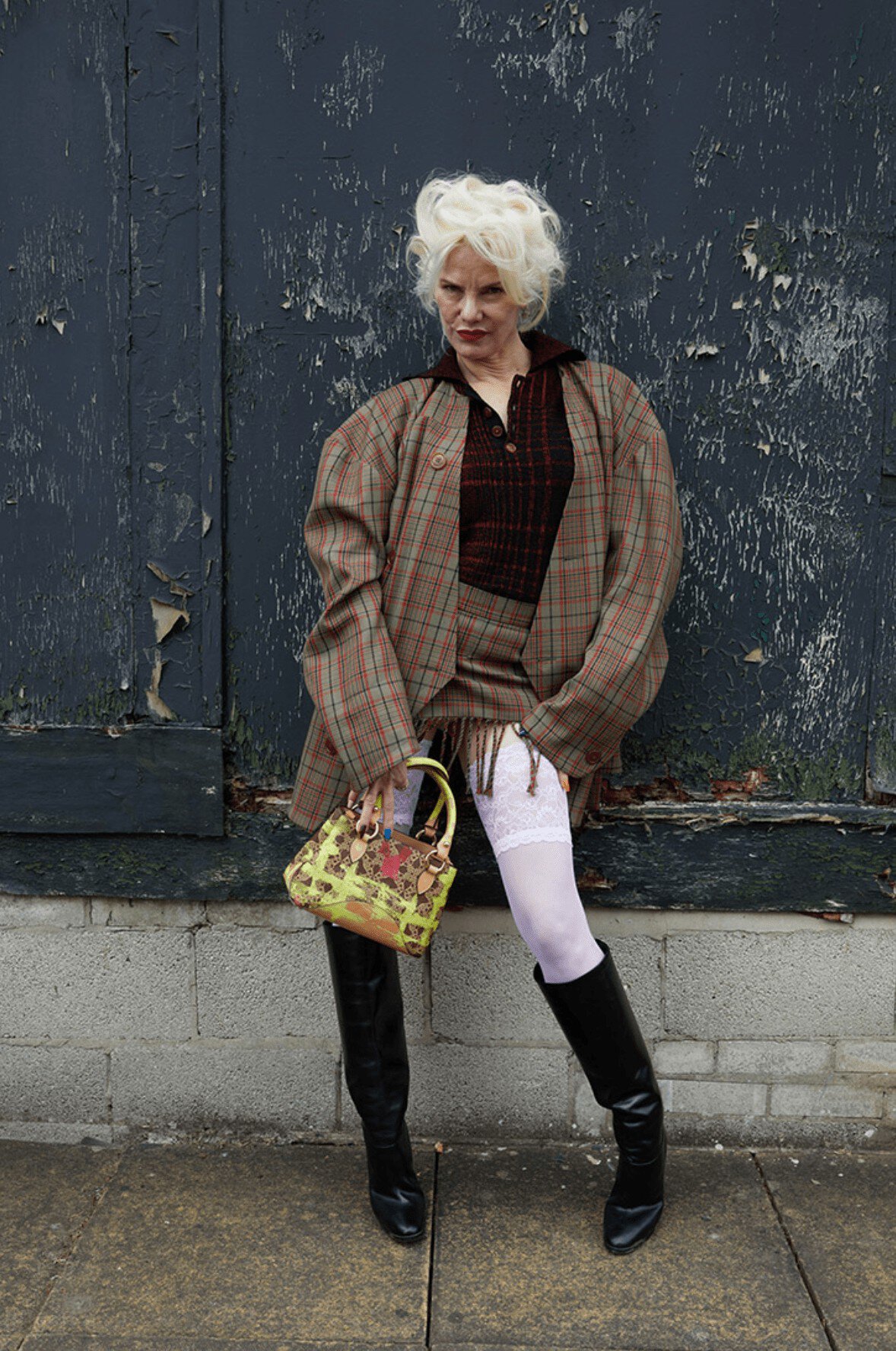Elegant mature woman in a plaid skirt suit and black boots poses against a distressed blue wall with a green handbag, fashion shot.