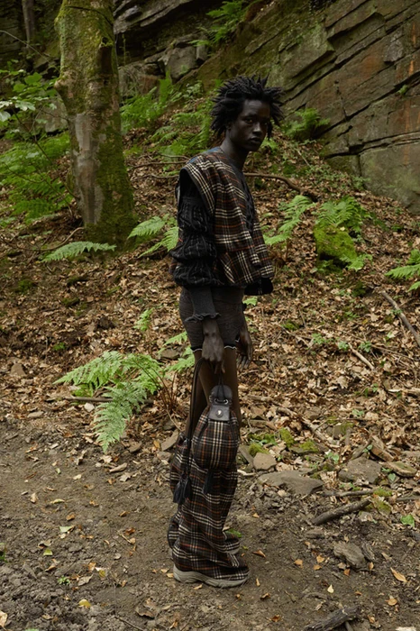 A model standing in a rocky woodland wearing a brown plaid vest, matching oversized plaid boots, and holding a bucket bag.