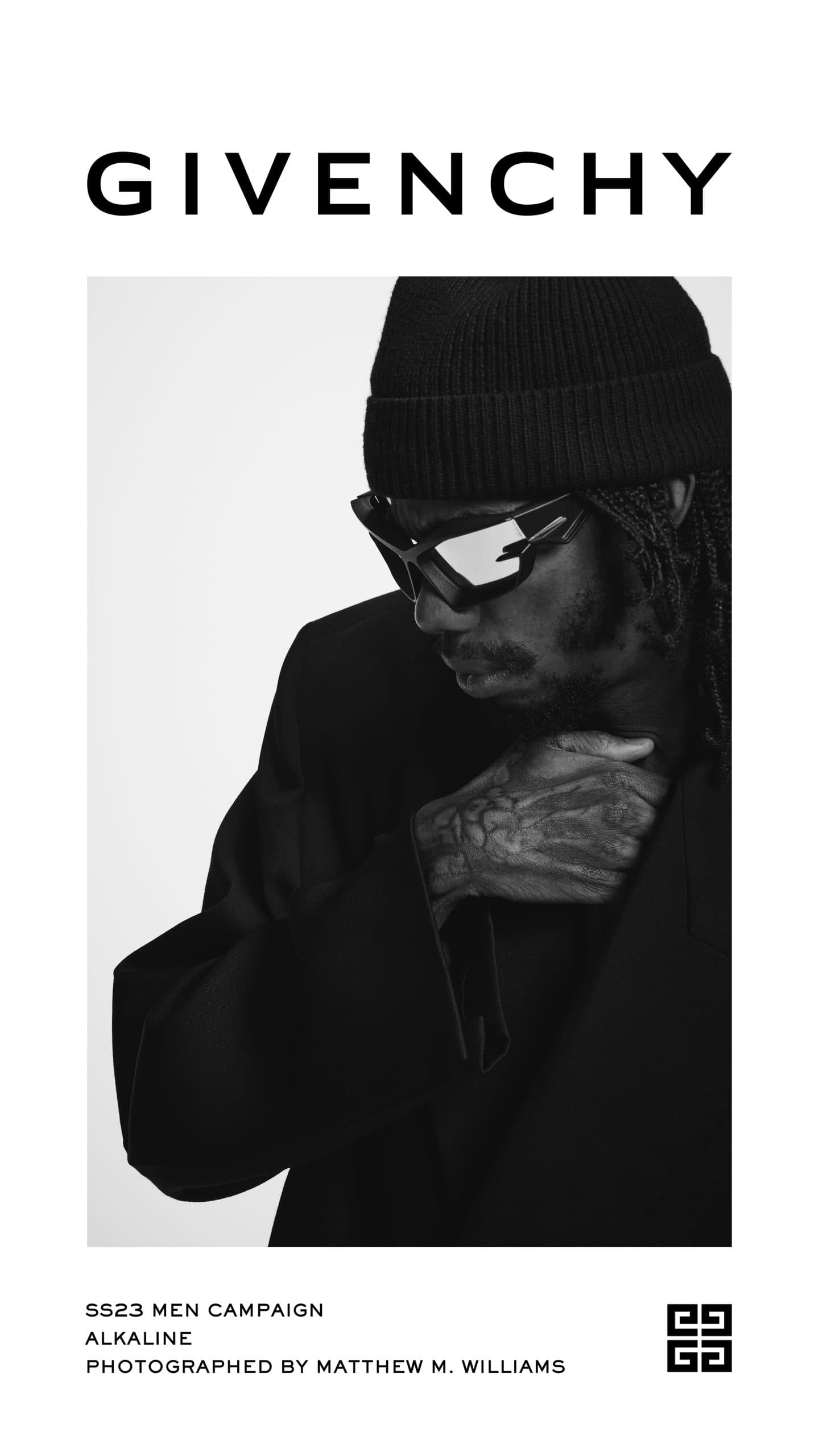 Black and white fashion campaign shot features a man in a beanie and sunglasses, wearing a dark blazer, against a light background.