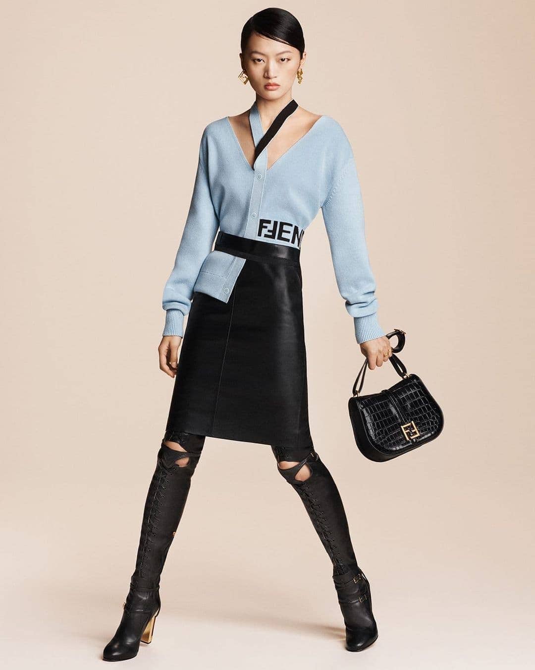An Asian female fashion model poses in a studio, showcasing a light blue V-neck sweater, black skirt, black boots, and a black handbag.