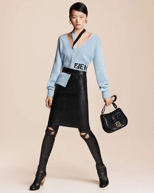 An Asian female fashion model poses in a studio, showcasing a light blue V-neck sweater, black skirt, black boots, and a black handbag.