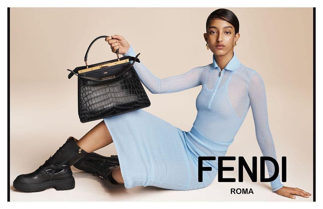 Fashion shot of a woman posing with a Fendi handbag and boots, wearing a light blue dress, in a studio setting against a neutral background.