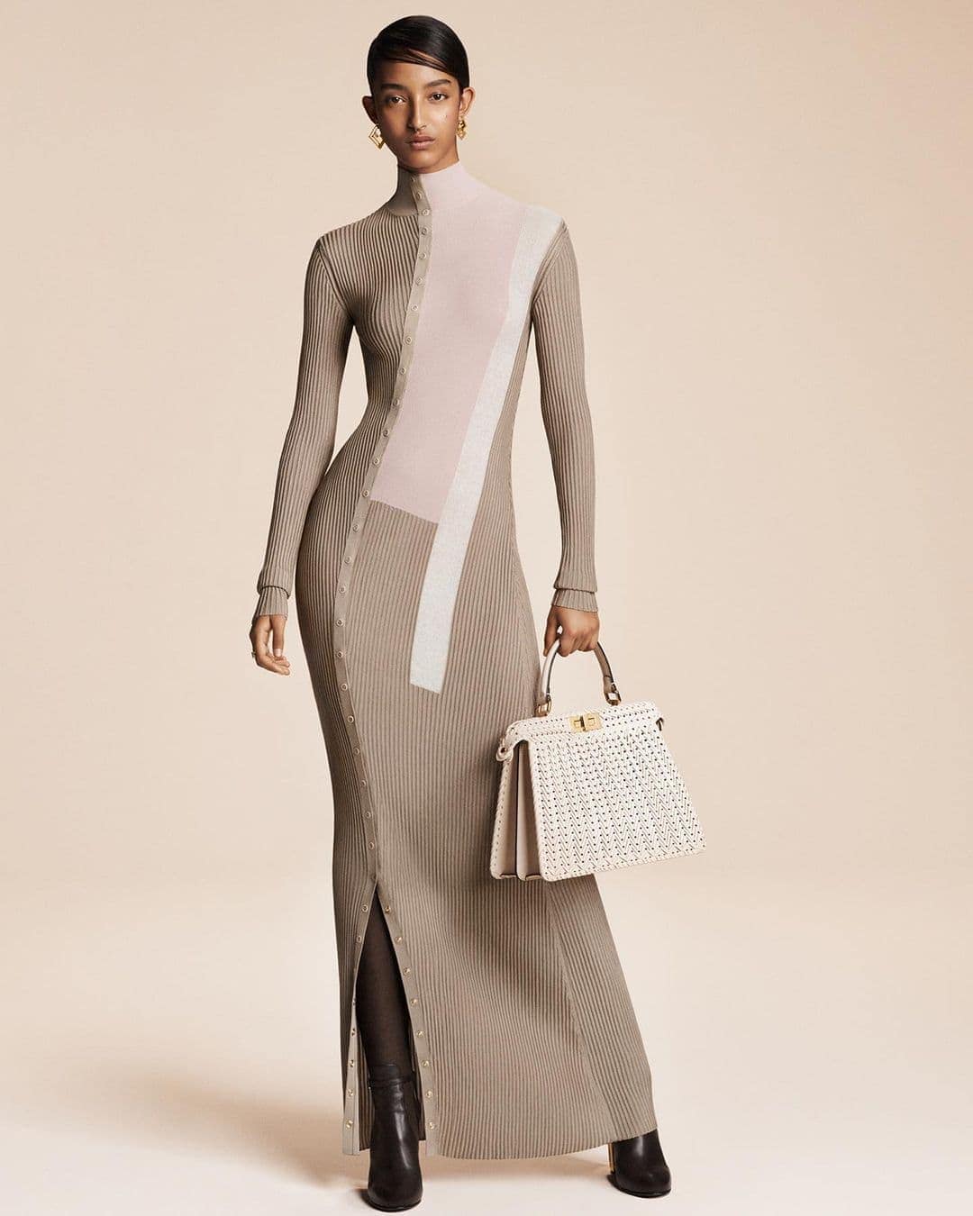 Elegant woman poses in a ribbed, long-sleeved maxi dress, holding a woven handbag against a neutral backdrop in a fashion editorial shot.