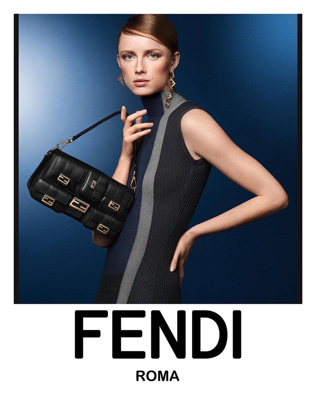 Fashion shot of a red-haired woman in a gray-and-black dress holding a black Fendi handbag against a deep blue background.