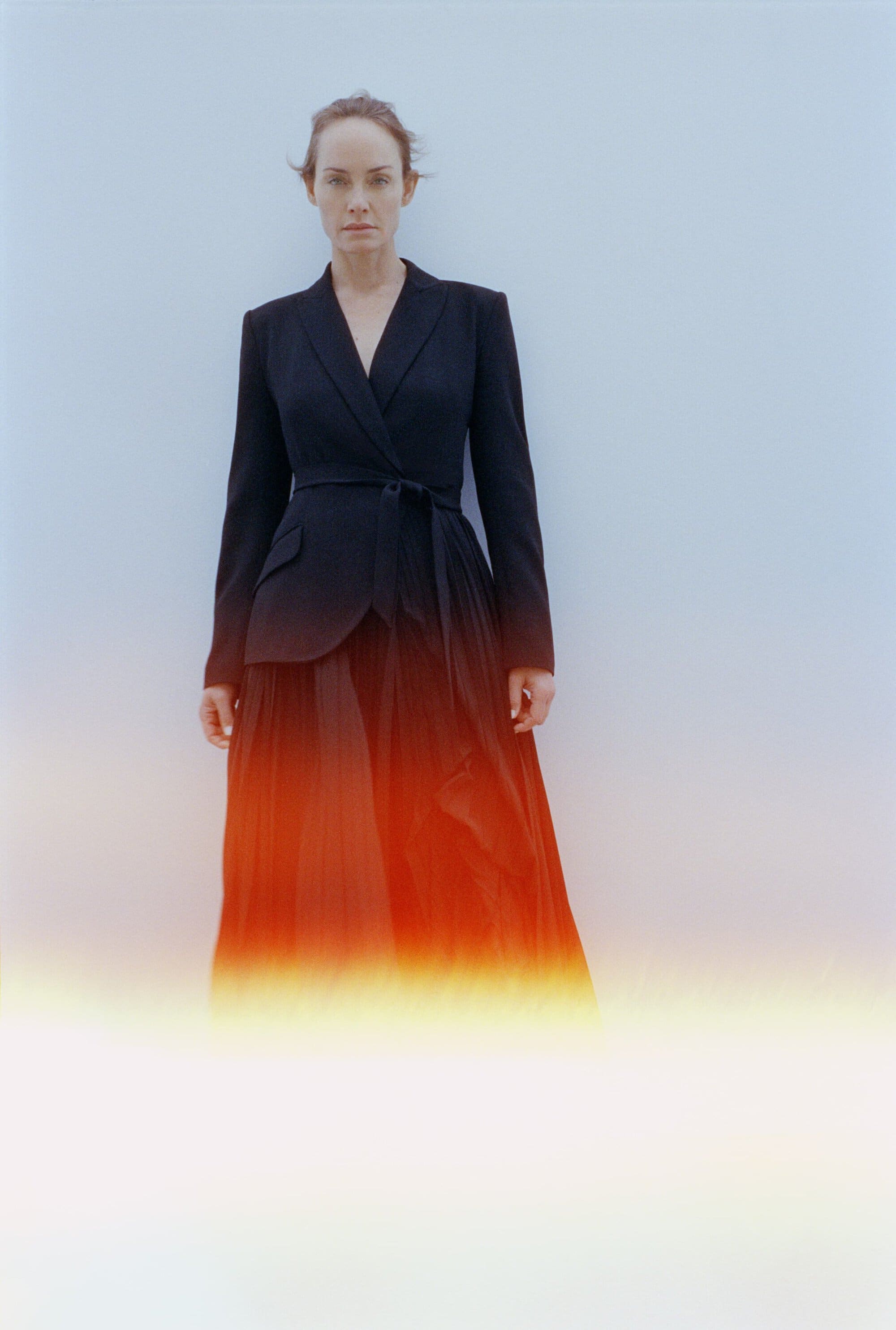 A woman poses in a black blazer and long black skirt against a light blue background, presented in a medium studio shot.