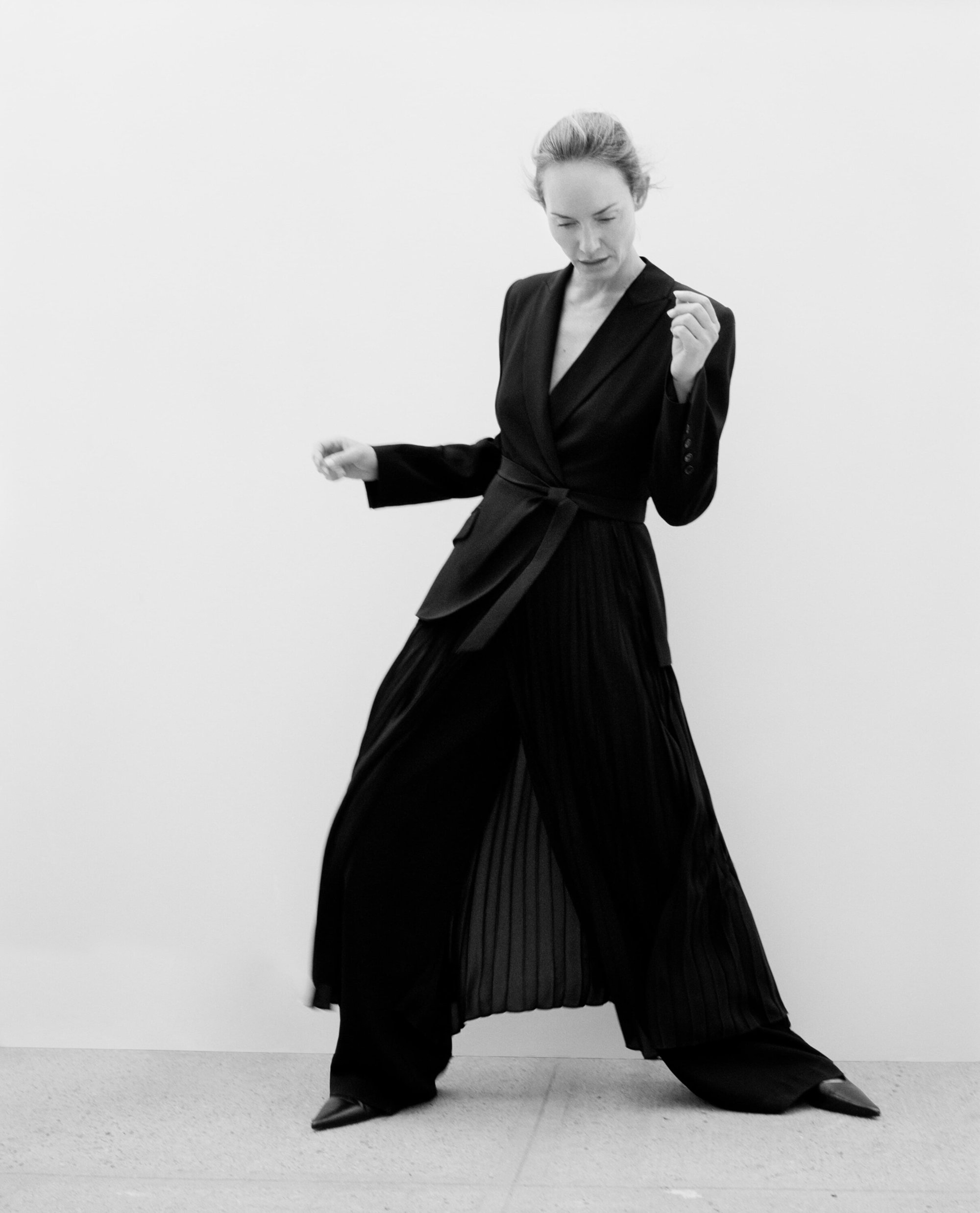 Elegant woman in black blazer and wide-leg trousers strikes a pose against a bright wall in this black and white fashion portrait.
