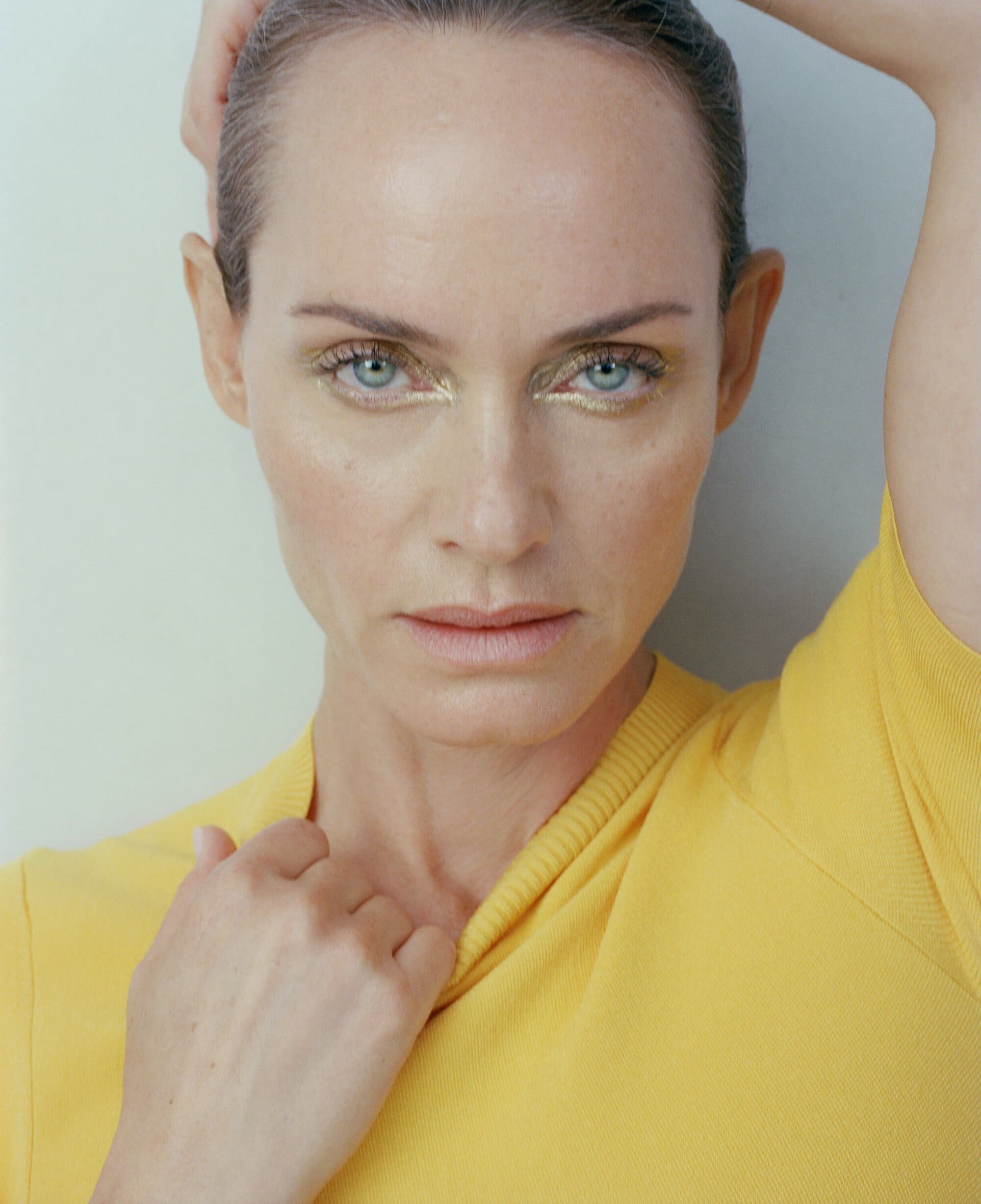 Close-up portrait of an adult female with golden eye makeup, wearing a yellow top, set against a neutral background.