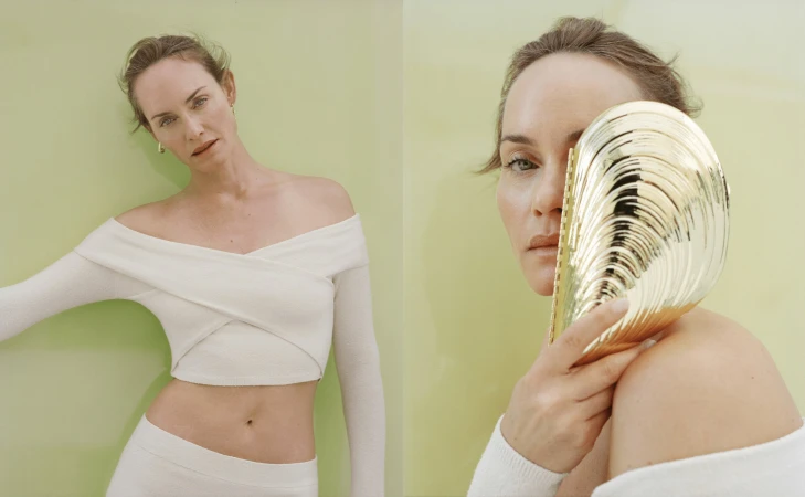 Studio shot of an adult woman in an off-the-shoulder crop top, posing with a gold seashell in front of a light green backdrop.
