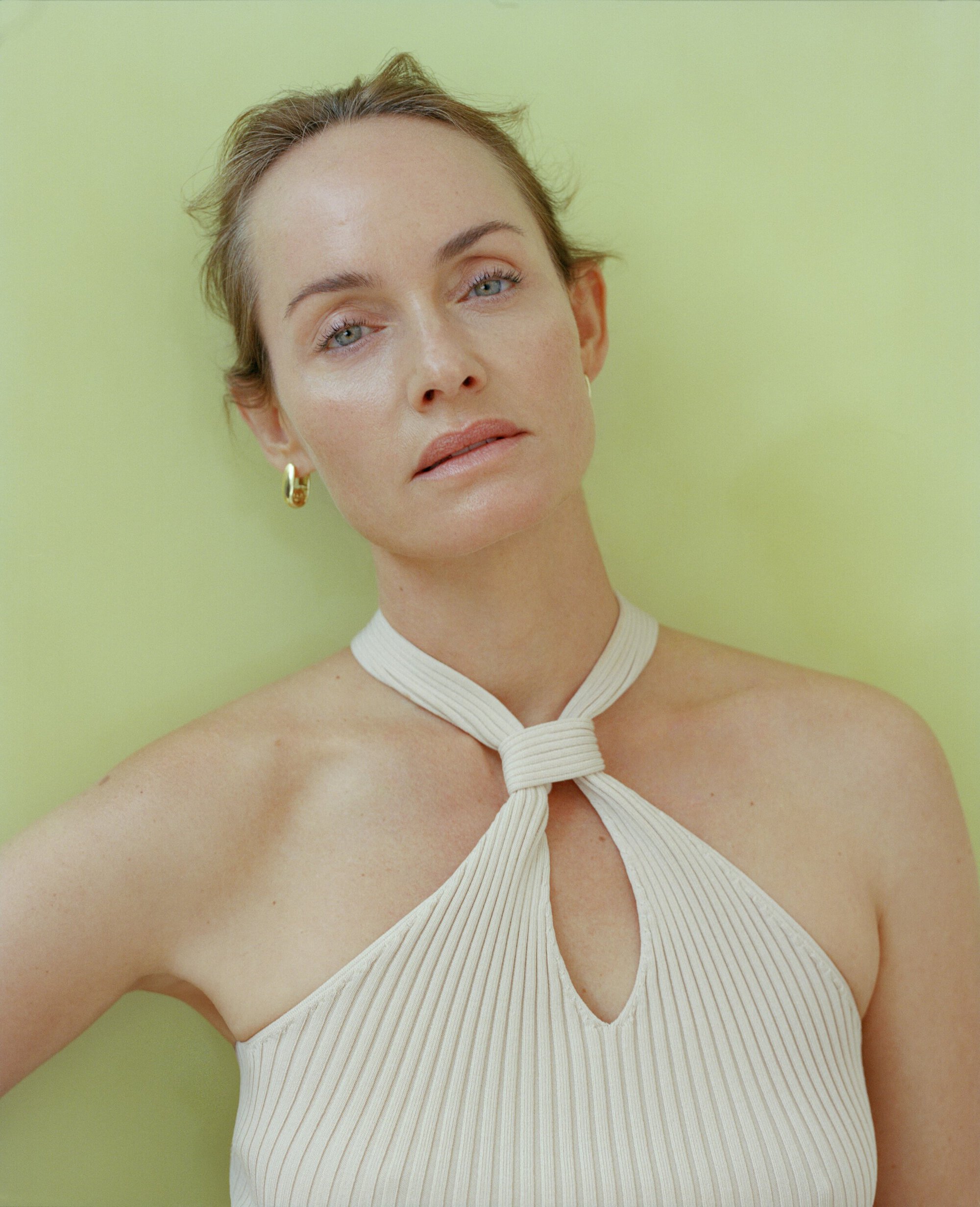 Close-up portrait of an adult woman with fair skin wearing a sleeveless, textured halter top against a solid light green background.