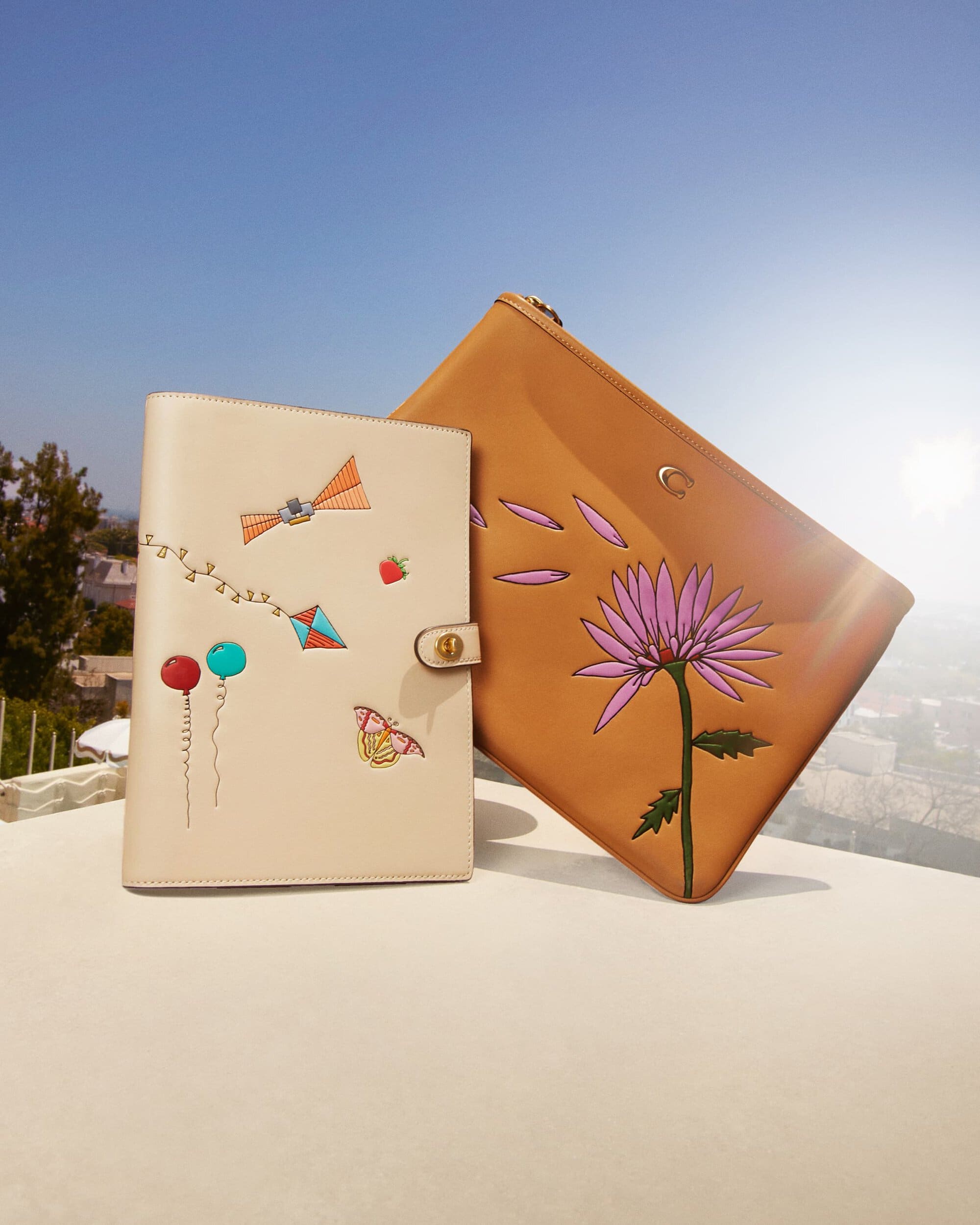 Tan-colored leather planner and pouch with hand-painted floral and kite designs, displayed outdoors against a blue sky. Fashion accessory.