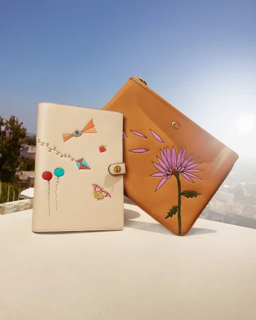 Tan-colored leather planner and pouch with hand-painted floral and kite designs, displayed outdoors against a blue sky. Fashion accessory.