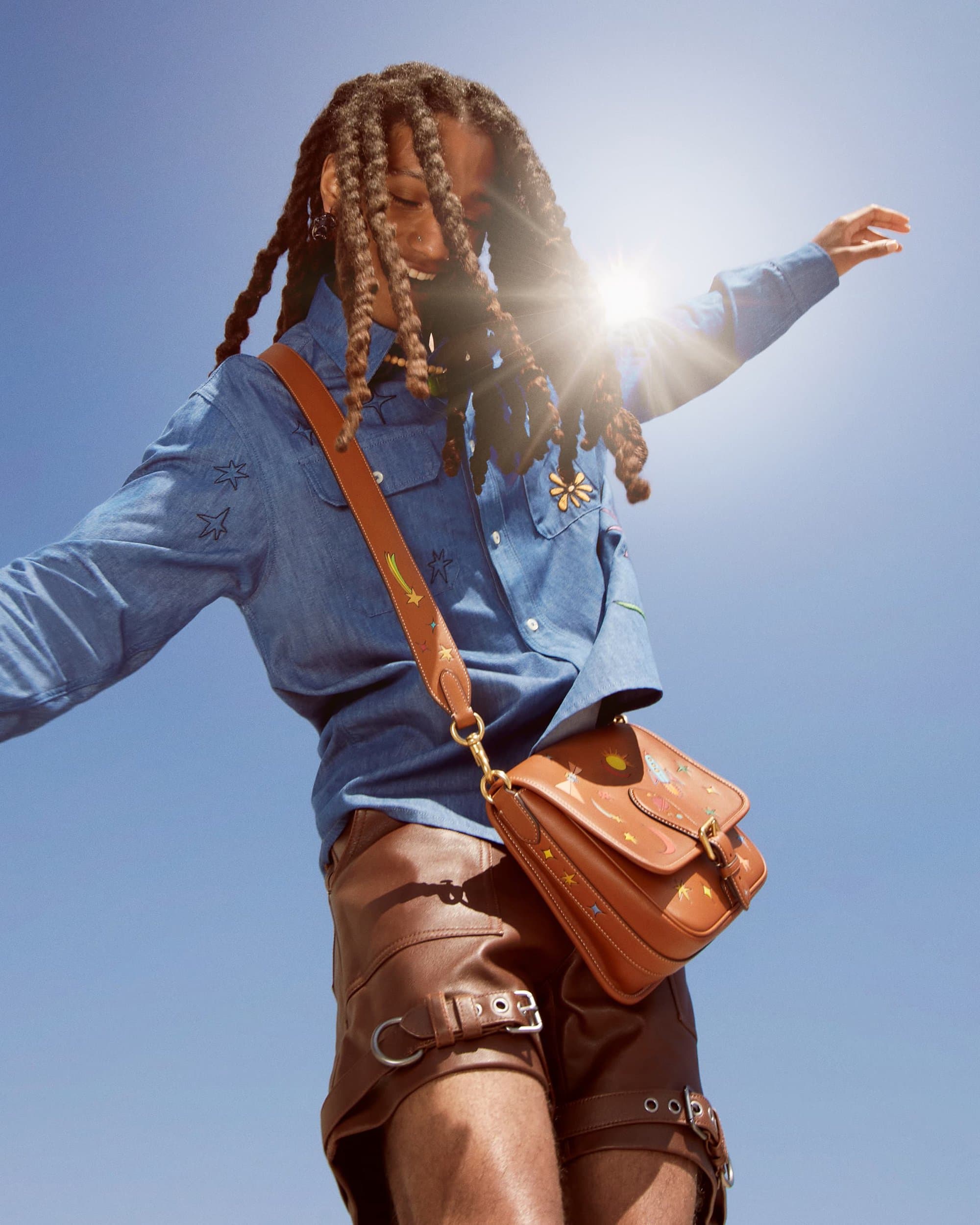 An adult with dreadlocks wearing a blue denim shirt, brown leather shorts, and a brown leather crossbody bag against a blue sky.