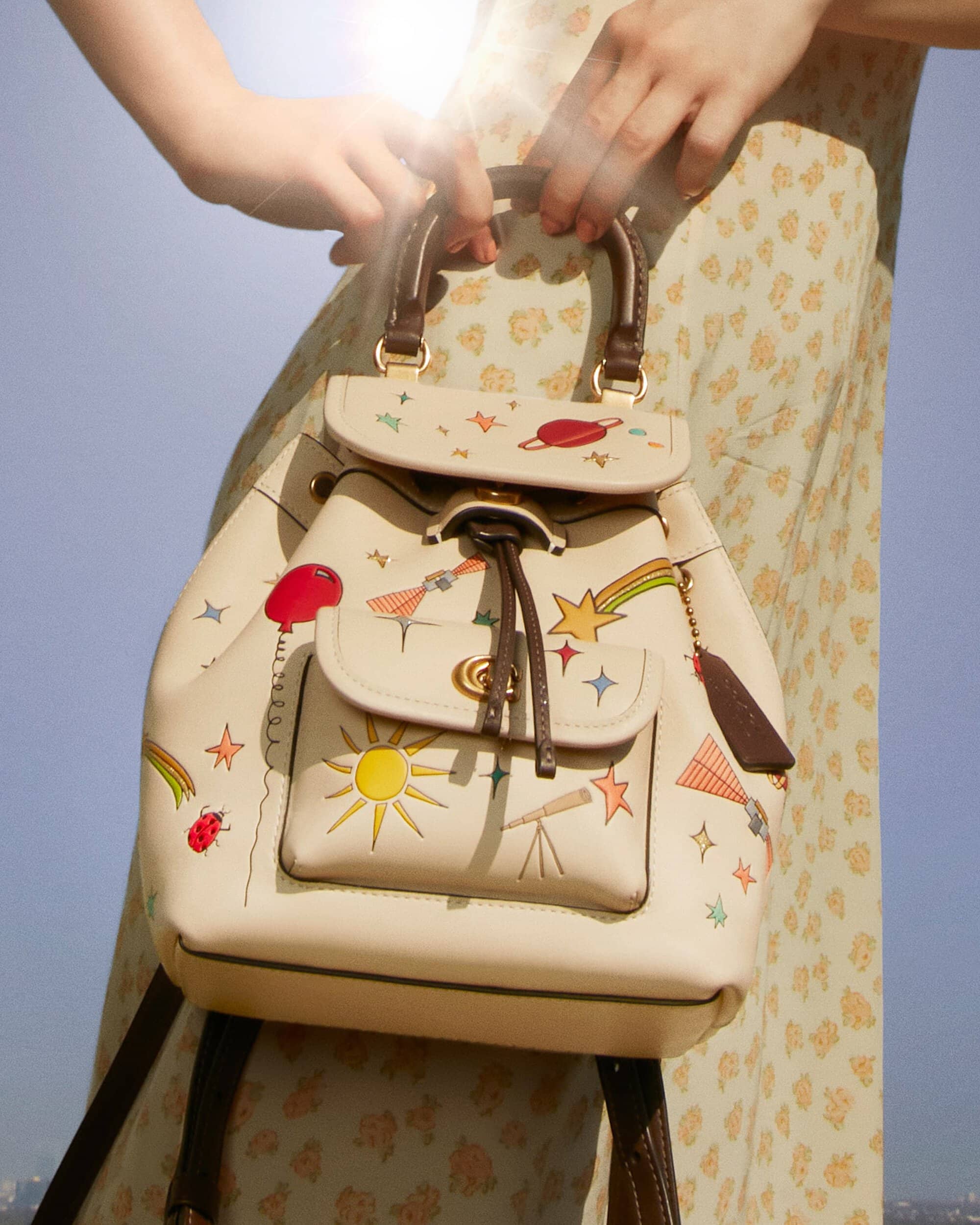 Close-up of a person holding a Coach backpack with cartoon-style space and celestial-themed designs; a woman in a floral-patterned dress stands behind.