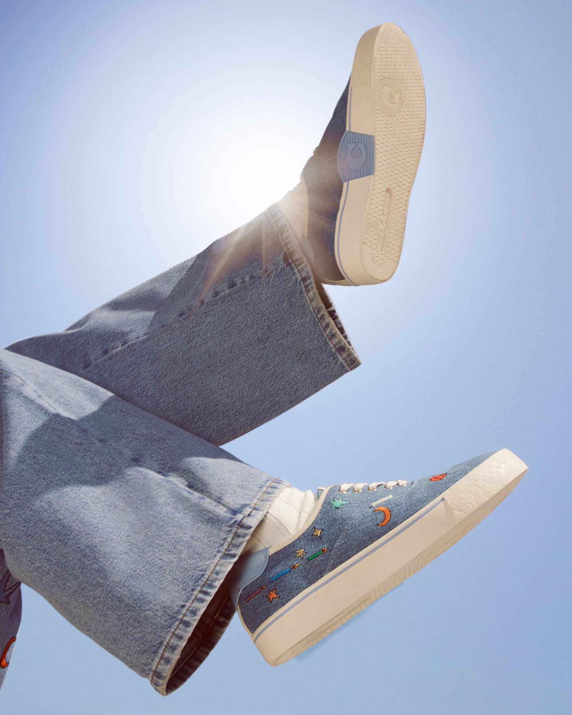 Elevated view of light-blue denim sneakers with embroidered stars and jeans against a bright-blue sky with sun glare.
