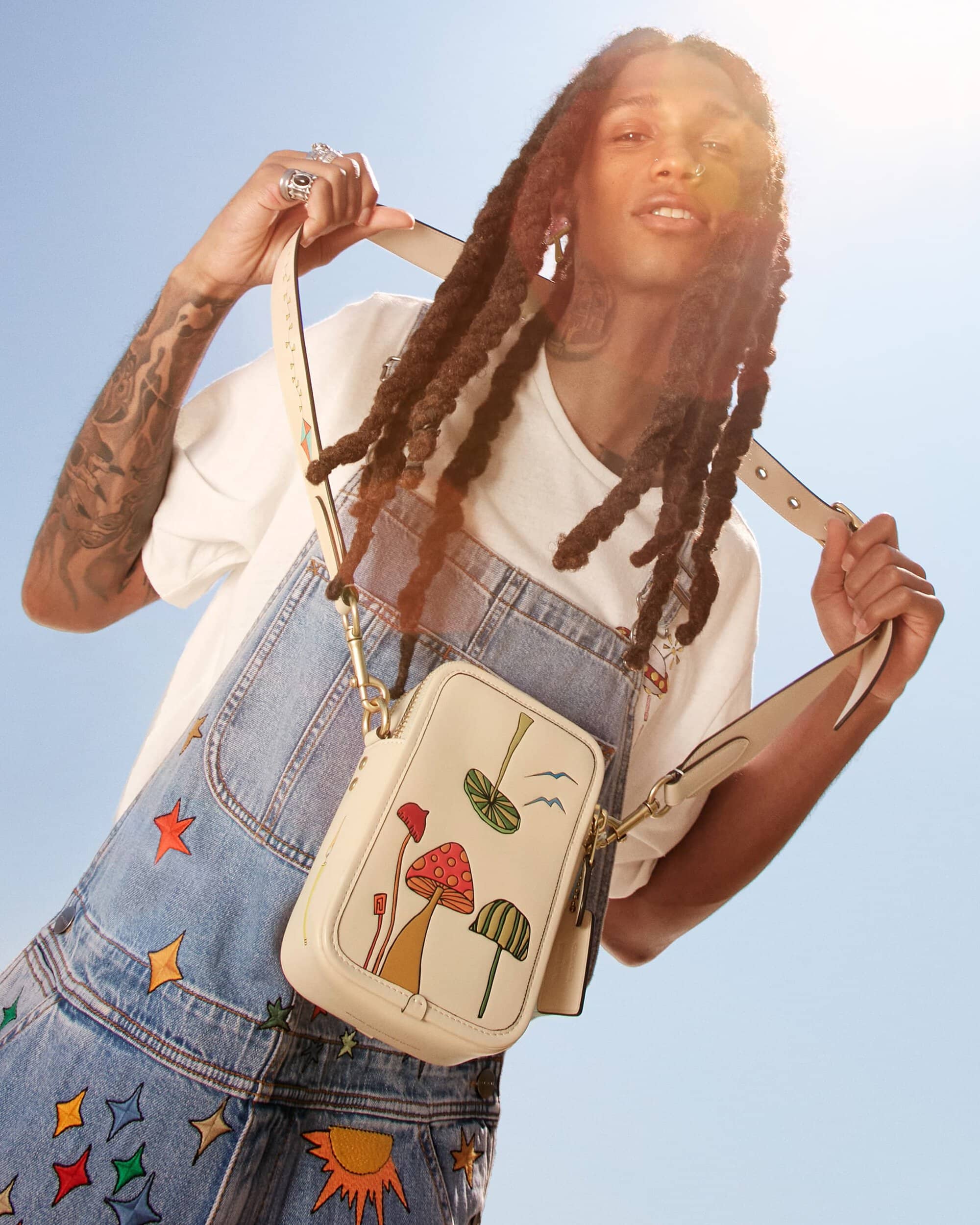 A person with dreadlocks and a white t-shirt wears a blue jean overall dress with colorful star designs, posing with a purse.