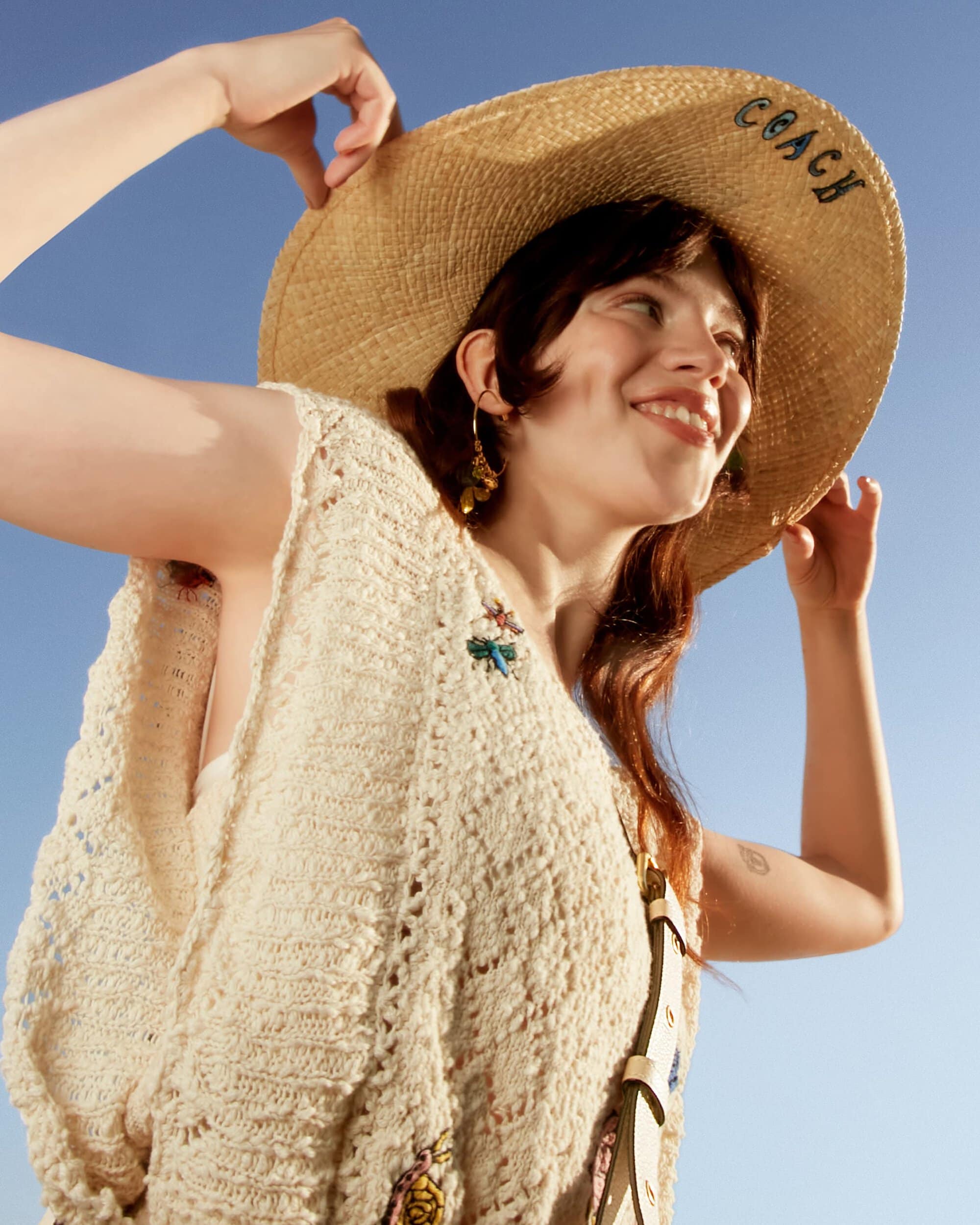 Woman in crochet top and straw hat smiles against blue sky. The hat bears the Coach logo. Close up shot, outdoor setting, eye-level angle.
